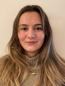 Portrait d'une jeune femme avec des cheveux longs et blonds, portant un pull beige et des colliers en or, de face contre un fond clair.