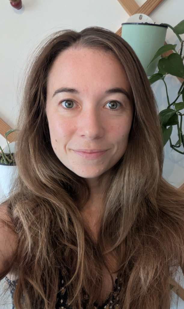 Portrait d'une femme souriante avec de longs cheveux bruns, prenant la photo à l'intérieur, entourée de plantes.